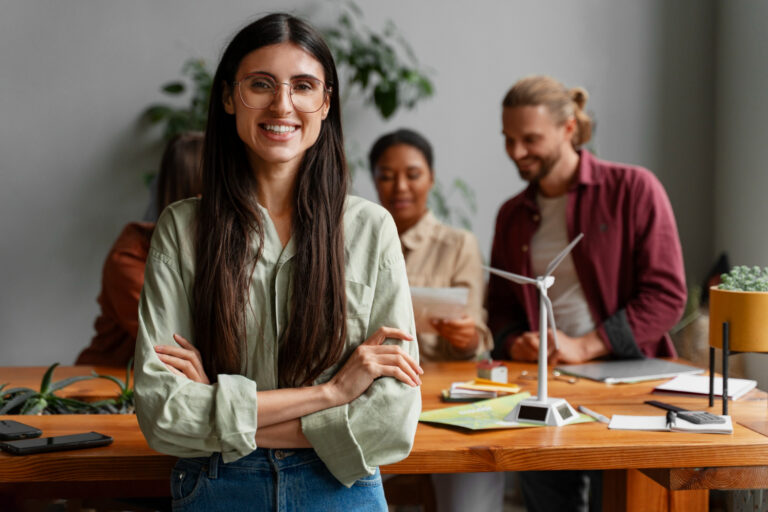 woman standing in front of desk, arms crossed and smiling with colleagues work in the background